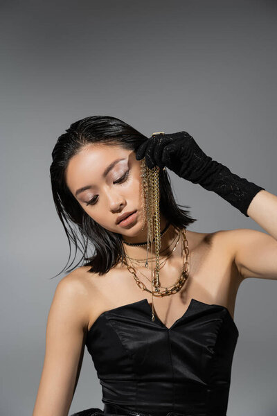 portrait of brunette and asian young woman with short hair posing in black gloves and strapless dress while holding golden jewelry on grey background, wet hairstyle, natural makeup
