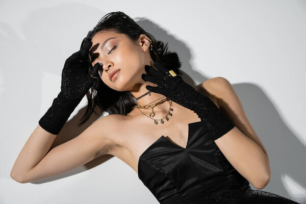 top view of young asian woman with closed eyes and short brunette hair lying in black gloves and strapless dress while posing in golden jewelry on grey background, wet hairstyle, hands near face 