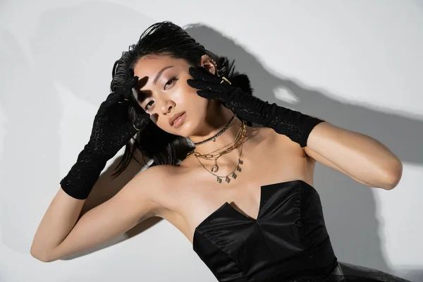top view of mesmerizing young asian woman with short brunette hair lying in black gloves and strapless dress while posing in golden jewelry on grey background, wet hairstyle, hands near face