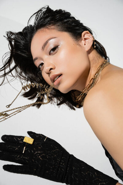 fashionable asian model with short brunette hair holding golden jewelry in mouth while looking at camera and lying on grey background, wet hairstyle, young woman, black gloves with rings 