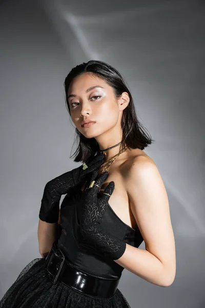 stock image portrait of alluring asian young woman with short hair posing in black strapless dress with belt and gloves while looking at camera on grey background, wet hairstyle, golden necklaces 