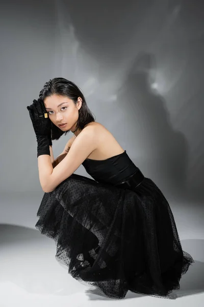 stock image full length of mesmerizing asian young woman with short hair sitting in black strapless dress with tulle skirt and gloves while looking at camera on grey background, wet hairstyle, golden necklaces 