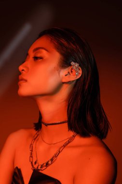 portrait of beautiful asian woman with short hair and wet hairstyle posing in strapless dress with trendy cuff earring and necklaces on dark orange background with red lighting, young model  clipart