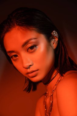 portrait of mesmerizing asian woman with short hair and wet hairstyle posing with trendy cuff earring and necklaces on dark orange background with red lighting, young model, looking at camera clipart