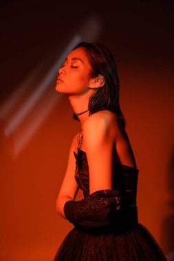portrait of stunning asian woman with short hair and wet hairstyle posing in strapless dress and glove with trendy cuff earring and necklaces on dark orange background with red lighting, young model  clipart