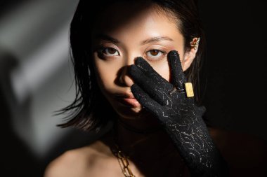 Asyalı genç bir kadının portresi, ıslak saç stili ve siyah eldivenli kısa saçlı, altın yüzüklü, gri arka planda duran, manken, kameraya bakan, gölgeler, koyu renk.