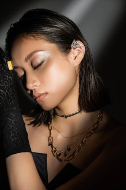 portrait of enchanting asian young woman with wet hairstyle and short hair posing in black glove with ring and ear cuff while standing on grey background, young model, closed eyes, shadows, dark clipart