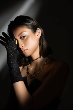 portrait of enchanting asian young woman with wet hairstyle and short hair posing in black glove with golden rings and ear cuff while standing on grey background, young model, shadows, dark clipart