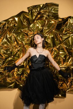 enchanting asian young woman with short hair posing in black strapless dress with tulle skirt and gloves while standing next to shiny yellow background, model, wrinkled golden foil, looking away clipart