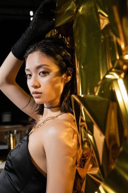 asian young woman with wet short hair posing in strapless dress with black glove and ear cuff while standing next to shiny background, model, looking at camera, wrinkled golden foil  clipart