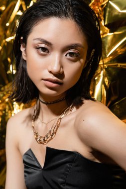 beautiful asian young woman with wet hairstyle and short hair posing in strapless dress with black glove while standing next to shiny golden background, model, looking at camera, wrinkled golden foil clipart