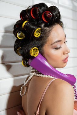 Saç bigudileri ve inci kolyeli şık ve Asyalı genç bir kadın mor retro telefonda beyaz fayansların yanında konuşuyor, ev hanımı, eski moda, eski moda. 