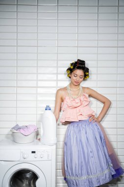 Saçları bukleli Asyalı genç bir kadın fırfırlı üst, inci kolye ve tül etek ile çamaşır odasında deterjanı olan, modern çamaşır makinesinde kirli giysileri ile çamaşır yıkama kabının yanında duruyor. 