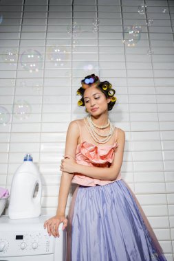 Saçları bukleli güzel Asyalı genç bir kadın fırfırlı üst, inci kolye ve tül etek içinde modern çamaşır makinesinin yanında duruyor çamaşır odasında deterjanı var, ev kadını, başka tarafa bakıyor, sabun köpükleri.