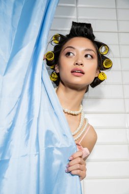 curious and asian young woman with hair curlers standing in pearl necklace behind blue bathroom curtain and looking away near white tiles at home, housewife, domestic scene  clipart