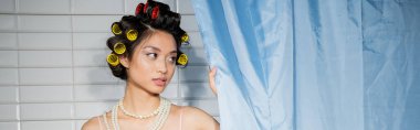 brunette and alluring asian woman with hair curlers standing in pearl necklace near blue bathroom curtain and looking away near white tiles at home, banner, housewife  clipart