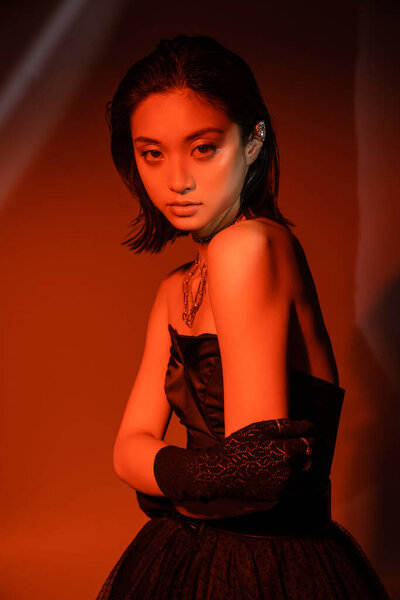 portrait of beautiful asian woman with short hair and wet hairstyle posing in strapless dress and glove with trendy cuff earring and necklaces on dark  orange background with red lighting 