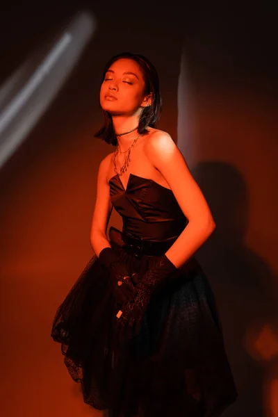 attractive asian woman with wet hairstyle posing in strapless dress with tulle skirt and black gloves with rings while standing on dark orange background with red lighting, young model, closed eyes