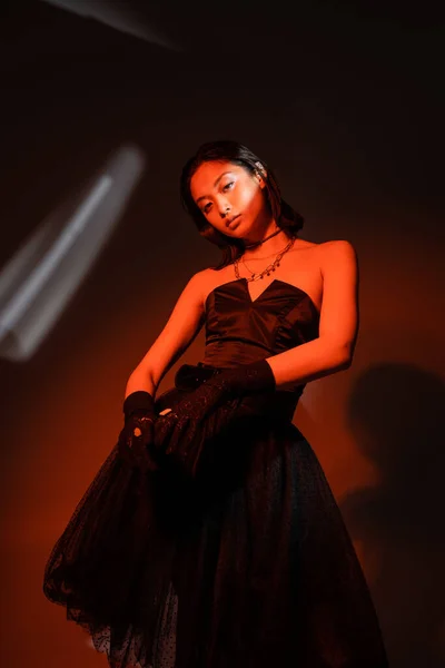 captivating asian woman with wet hairstyle posing in strapless dress with tulle skirt and black gloves with rings while standing on dark orange background with red lighting, looking at camera