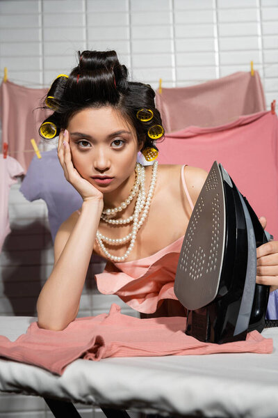 bored asian housewife with hair curlers holding iron while posing in pink ruffled top and pearl necklace near clean and wet clothes hanging on blurred background, housework, young woman, laundry 