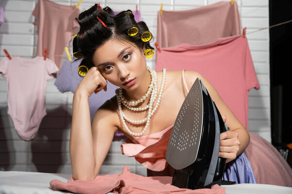 tired asian housewife with hair curlers in pink ruffled top and pearl necklace looking at camera while holding iron near clean clothes hanging on blurred background, housework, young woman, laundry 