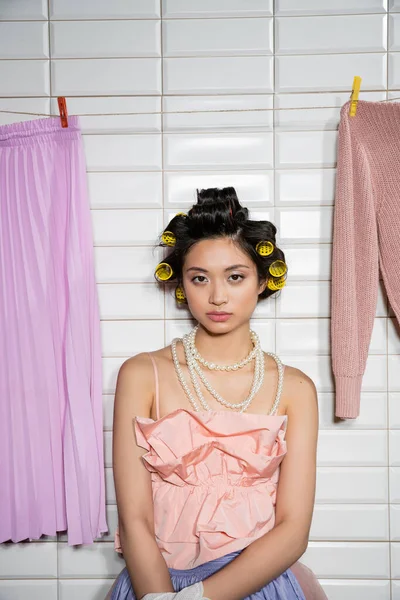 ピンクのフリルの上に立っている髪のカールを持つアジアの若い女性とカメラを見て 白のタイル 主婦の近くにぶら下がっている清潔で湿った洗濯真珠のネックレス — ストック写真