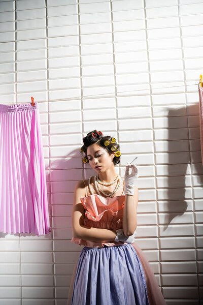 азиатская молодая женщина с бигуди волос стоя в розовом потрепанный топ, жемчужное ожерелье и перчатки, держа сигарету возле мокрой прачечной висит рядом с белой плиткой, домохозяйка, курение, плохая привычка 