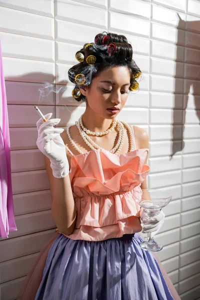 ピンク色のフリルの上に立って髪のカールを持つアジアの若い女性 濡れた洗濯や白いタイルの近くにタバコやガラスを保持しながら真珠のネックレスと手袋 — ストック写真