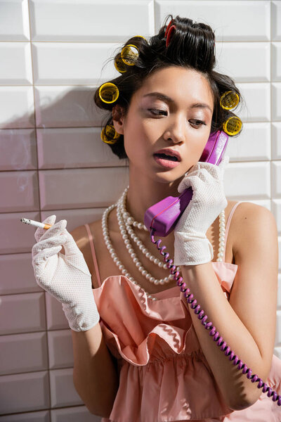 брюнетка и азиатская молодая женщина с бигуди волос, стоящие в розовой рифленой верхней части, жемчужное ожерелье и белые перчатки курить и говорить по ретро-телефону рядом с белой плиткой, домохозяйка, держа сигарету 