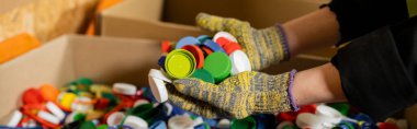 Çöp öğütme istasyonunda, çöp ayıklama ve geri dönüşüm konseptinde çalışırken, bulanık kutuların yanında renkli plastik kapaklar tutan koruyucu eldivenler giymiş kadın tasnif edicisi, afiş 