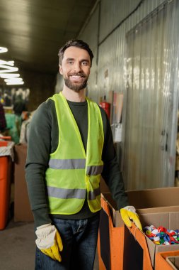 Yüksek görüş açısına sahip yelek ve koruyucu eldivenler giyen neşeli ayırıcı, çöp öğütme istasyonunda plastik kapsüllerin yanında dururken kameraya bakıyor, çöp ayıklama ve geri dönüşüm konsepti