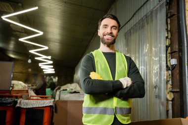 Yüksek görüş yeleği ve koruyucu eldivenleri olan neşeli işçiler kameraya bakıyor ve kollarını kavuşturuyor. Bulanık atık öğütme istasyonunda duruyor. Çöp ayıklama ve geri dönüşüm konsepti.