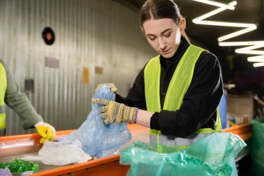 Eldivenli ve koruyucu elbiseli genç işçiler atık öğütme istasyonu, çöp ayıklama ve geri dönüşüm konseptinde çalışırken plastik torba ve taşıyıcının yanında çöp tutuyorlar.