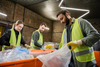 Koruyucu yelek ve eldiven giyen Hintli işçiler, bulanık iş arkadaşları ve atık öğütme istasyonunda, çöp ayıklama ve geri dönüşüm konseptinde çalışırken plastik çöpleri poşete koyuyorlar.