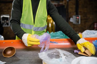 Cropped view of sorter in protective gloves and vest taking garbage from conveyor while working in blurred garbage sorting center, garbage sorting and recycling concept clipart