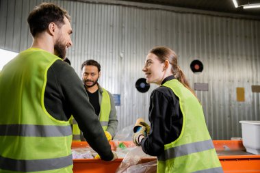 Koruyucu üniformalı ve eldivenli gülümseyen kadın çöpleri tutuyor ve atık öğütme istasyonunda birlikte çalışırken ırklar arası iş arkadaşlarıyla konuşuyor.