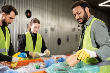 Bearded indian worker in safety vest and protective gloves putting paper cup in plastic bag while separating trash near conveyor and colleagues in waste disposal station, recycling concept clipart