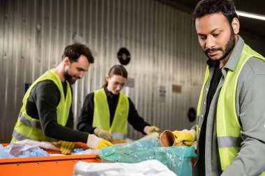 Eldiven ve yüksek görüş açısına sahip bir Hintli sakallı ayıklayıcı konveyör yanında çalışırken plastik torbaya kağıt bardak koyuyor ve atık imha istasyonunda bulanık meslektaşlar, geri dönüşüm konsepti