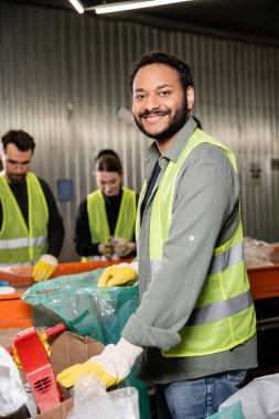 Koruyucu eldivenler ve güvenlik yeleği giymiş neşeli Hintli ayırıcı plastik torba yanında dururken kameraya bakıyor, bulanık taşıyıcı ve atık imha istasyonunda çalışan meslektaşlar, geri dönüşüm konsepti