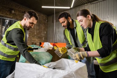 Koruyucu eldivenler ve güvenlik yelekleriyle ırklar arası sıralayıcılar çöp öğütme istasyonunda çöpleri ayıklarken plastik kapları torbadan çıkarıyorlar. Çöp ayıklama ve geri dönüşüm konsepti.