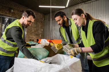 Interracial sorters in protective gloves and safety vests taking plastic containers from sacks while sorting trash together in waste disposal station, garbage sorting and recycling concept clipart
