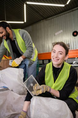Smiling young worker in reflective vest and gloves looking at camera while holding plastic garbage for recycle near sack and blurred indian colleague in waste disposal station, garbage sorting  clipart