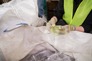 Cropped view of worker in protective gloves and safety vest holding plastic trash near sack for recycle while working in waste disposal station, garbage sorting and recycling concept clipart