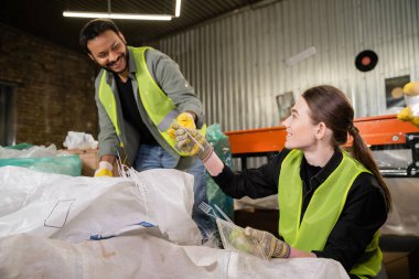 Bulanık atık öğütme istasyonunda, çöp ayıklama ve geri dönüşüm konseptinde çalışırken eldivenli ve koruyucu yelek giyen gülümseyen genç sıralayıcı kızılderili meslektaşına plastik çöp veriyor.