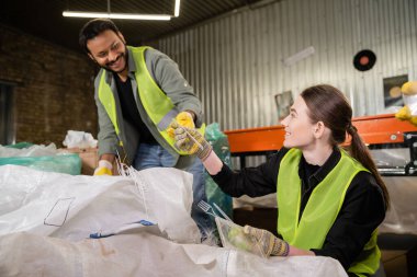 Smiling young sorter in gloves and protective vest giving plastic garbage to blurred indian colleague while working near sack in blurred waste disposal station, garbage sorting and recycling concept clipart