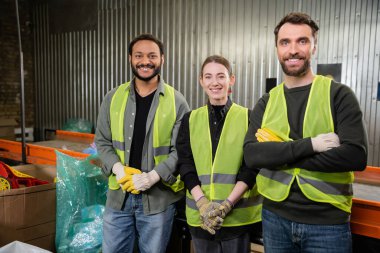 Positive interracial workers in high visibility vests and gloves looking at camera while standing near conveyor and working in waste disposal station, recycling concept clipart