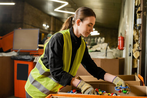 Young female worker of garbage sorting center wearing protective clothing and gloves while working with plastic caps in carton boxes, garbage sorting and recycling concept