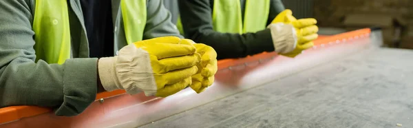Cropped view of sorter in protective gloves and vest standing near conveyor and blurred colleague while working in garbage sorting center, garbage sorting and recycling concept, banner