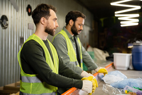 Side view of male sorter in high visibility vest and gloves standing near garbage on conveyor and blurred indian colleague in waste disposal station, garbage sorting and recycling concept