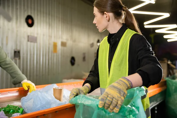 Koruyucu eldivenler ve yelek giyen genç işçiler çöp öğütme istasyonu, çöp ayıklama ve geri dönüşüm konseptinde çalışırken konveyörün yanında dururken plastik poşet tutuyorlar.